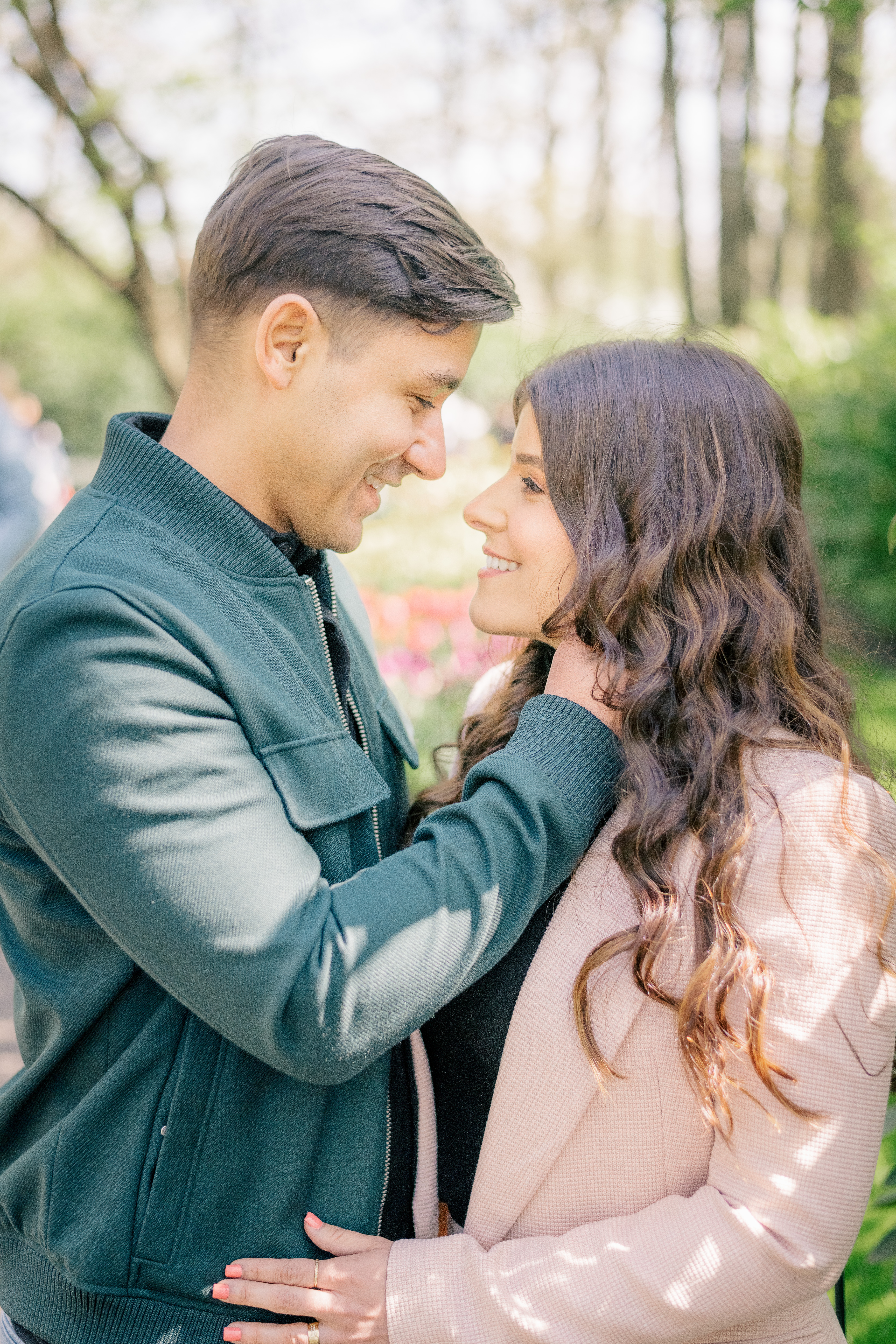 A woman and a man stand facing each other. The man holds the woman's head in his hands and they gaze into each other's eyes while standing in a garden