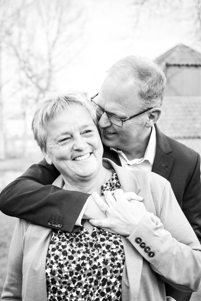 A man stands behind a woman with his arm around her shoulder. She takes his hand in her hand and they are both smiling